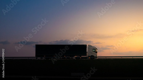 Silhouette of a truck on road at sunset.Semi truck trailer silhouette with setting sun.Delivery truck on asphalt road highway at sunset.Truck passing by a railing. Underexposed pic.