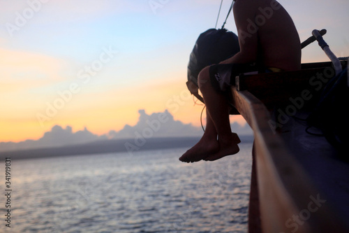 A young man sitting on the deck of a sailboat at sunrise, the man is pointing his finger towards the horizon. Komodo National Park. Indonesia