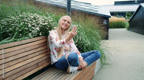 Woman making video call on green roof. Lovely young female using smartphone to make video call while sitting on bench on living roof of huge mall in USA. © art24pro