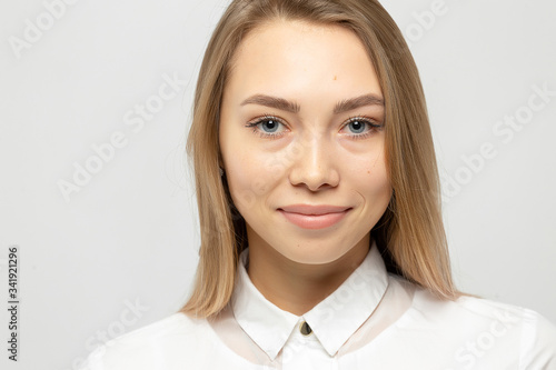 Close-up portrait of beautiful happy young attractive blonde woman in a white shirt with clean skin looking at camera against gray-white background.