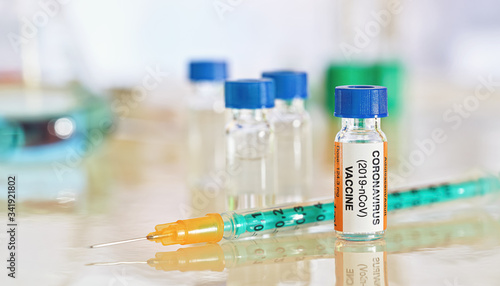 Coronavirus covid-19 vaccine concept - small bottle with blue cap on glossy white desk, green orange syringe near, more lab glass blurred background (sticker is own design - not real product)