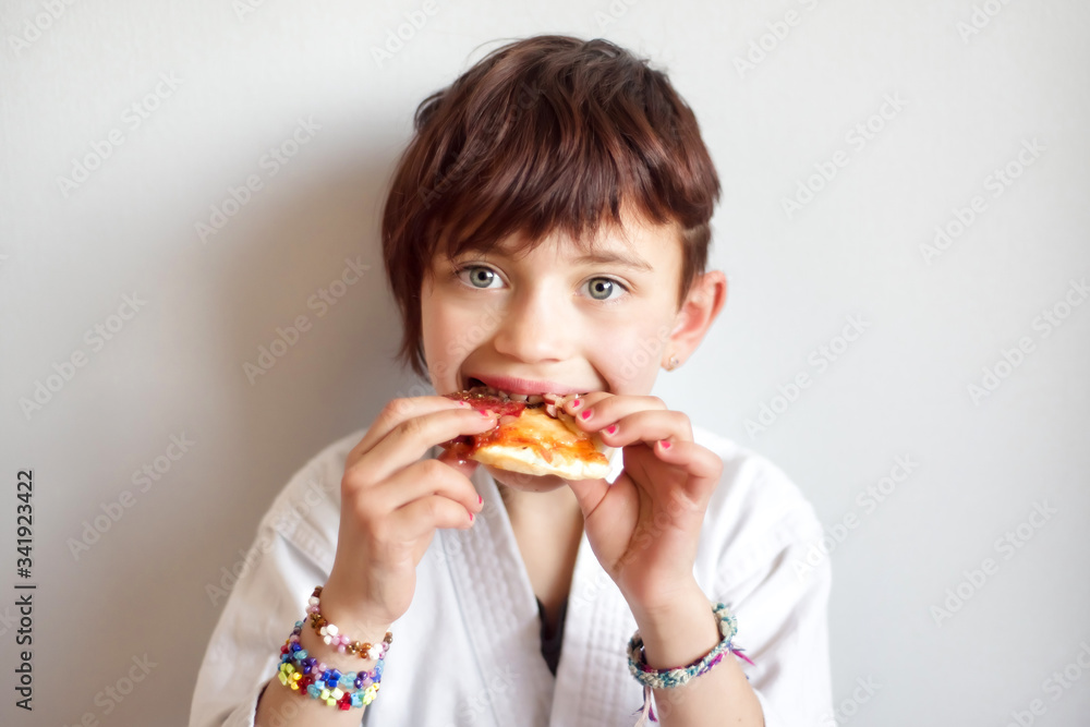 Portrait of cute 7 years old hungry short haired girl in white kimono eating pizza after hard on-line karate taekwondo training. Child after distance training eat fast food at home. Martial arts