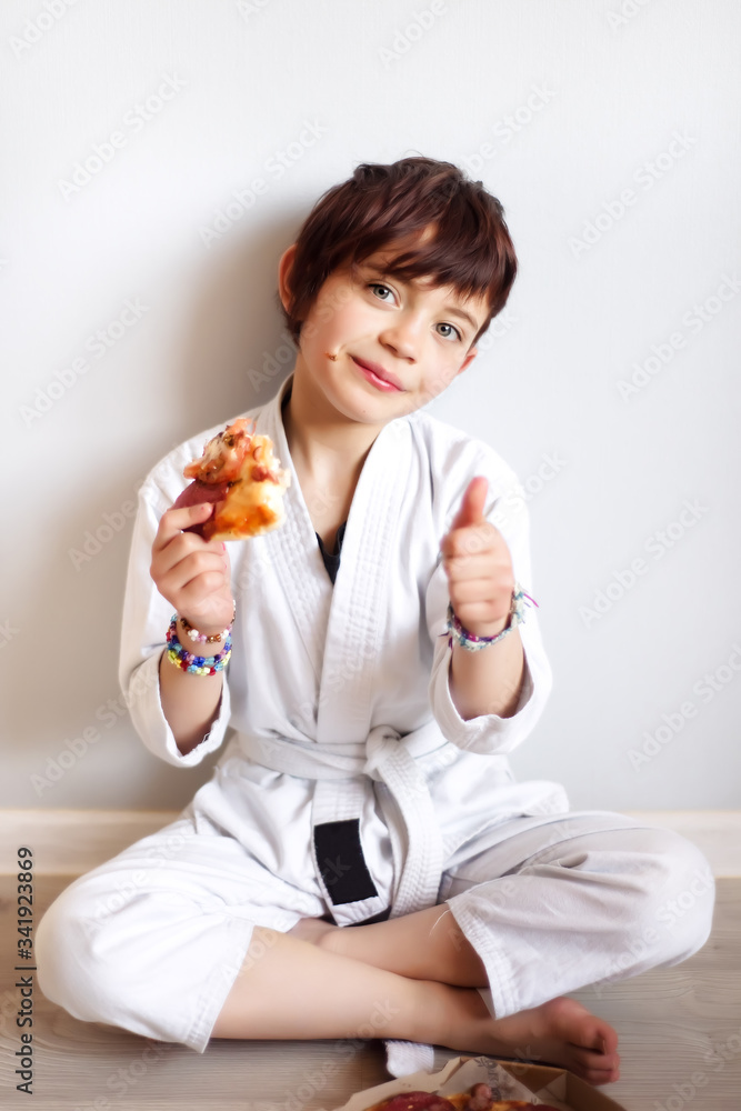 Vertical portrait of cute 7 years old hungry short haired girl in white kimono eating pizza after hard on-line karate taekwondo training. Child after distance training eat at home. Martial arts Vertical portrait of cute 7 years old hungry short haired girl in white kimono eating pizza after hard on-line karate taekwondo training. Child after distance training eat at home. Martial arts