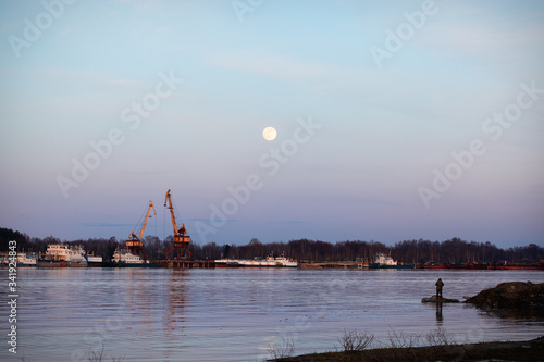 A man fishing in the evening from the bank of the Volga river against the backdrop of the rising moon and industrial facilities of the river port on the opposite bank