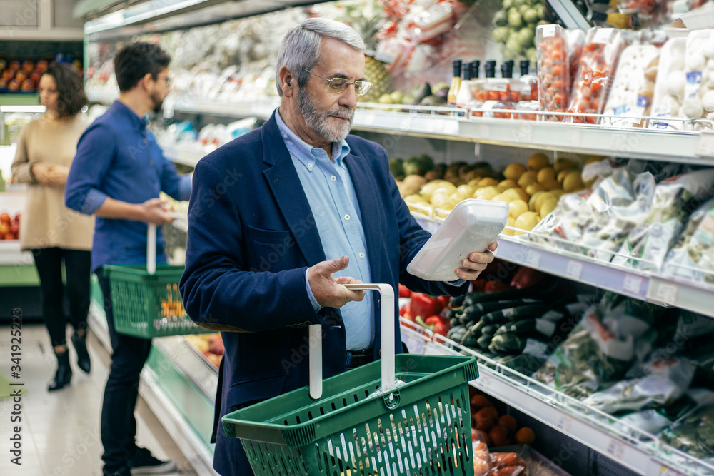 Mature man choosing vegetables in supermarket. Bearded man in eyeglasses holding basket and buying fresh vegetables in grocery store. Shopping concept