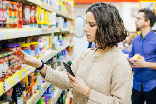 Woman holding smartphone and choosing goods in supermarket. Young woman reading checklist via smartphone and choosing products in grocery store. Supermarket concept