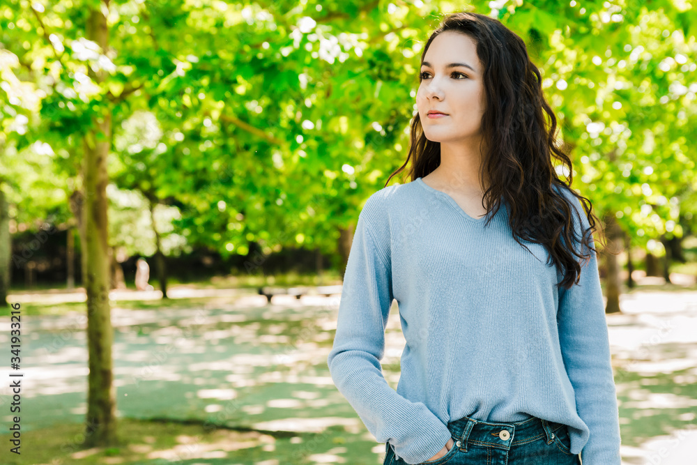 Beautiful woman standing in spring park and looking aside. Attractive young lady with long hair holding hand in pocket while posing in spring park. Female beauty concept Beautiful woman standing in spring park and looking aside. Attractive young lady with long hair holding hand in pocket while posing in spring park. Female beauty concept