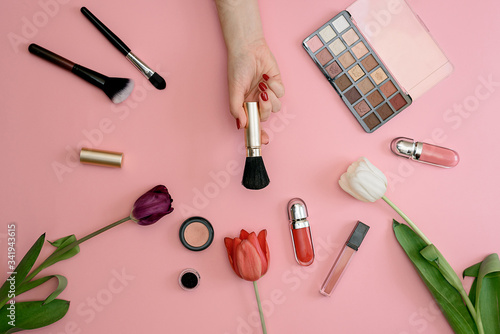 Girl holds makeup brush set of cosmetics and flowers on pink background