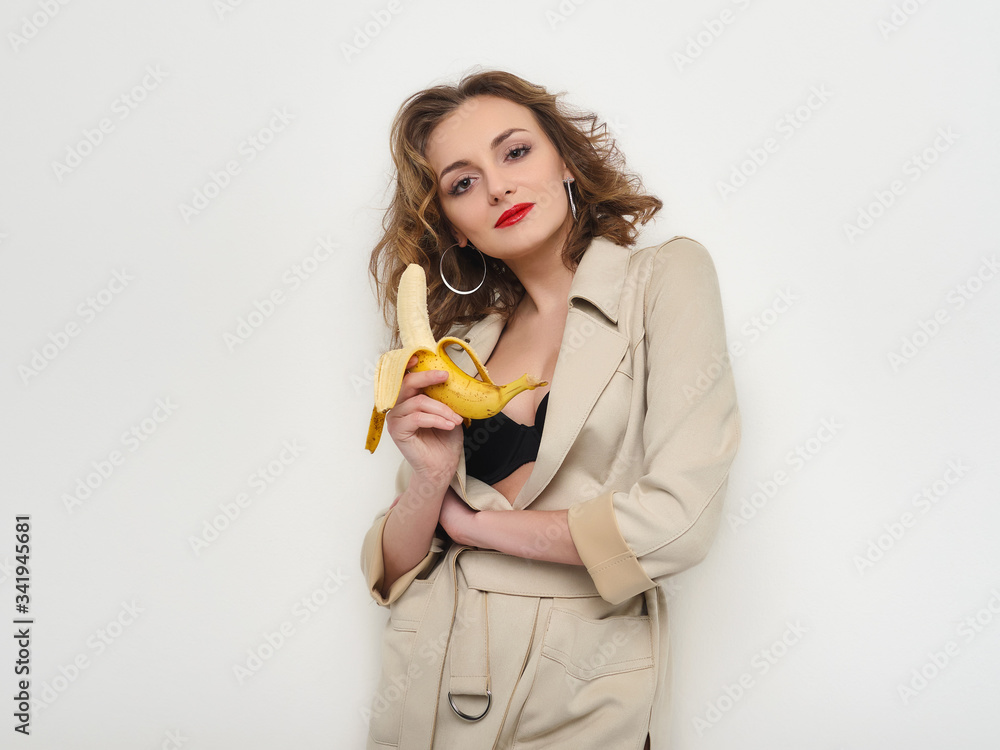 Sexy woman with red lips and curly hairdo in short coat and black bra playfully posing eating banana on white background Sexy woman with red lips and curly hairdo in short coat and black bra playfully posing eating banana on white background