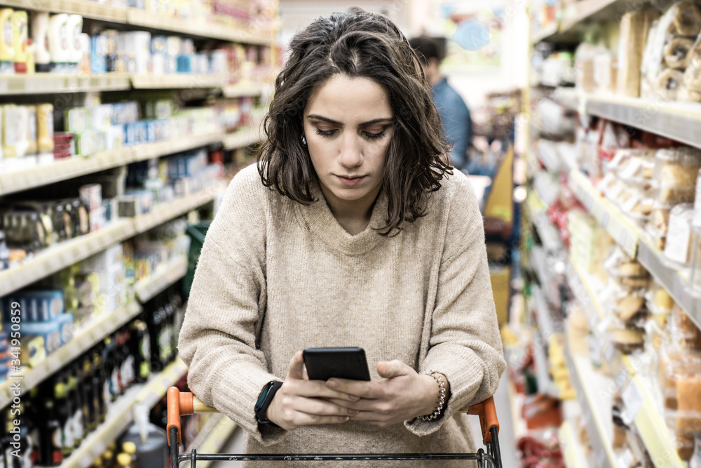 Woman leaning at cart and using smartphone in store. Young woman reading checklist via smartphone and choosing products in grocery store. Supermarket concept