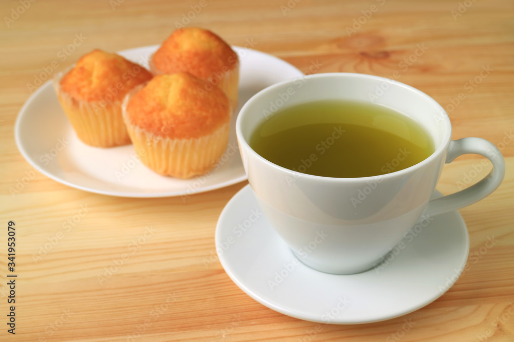 Cup of Hot Green Tea with Blurry Madeleine Cupcakes in Background Served on Wooden Table Cup of Hot Green Tea with Blurry Madeleine Cupcakes in Background Served on Wooden Table