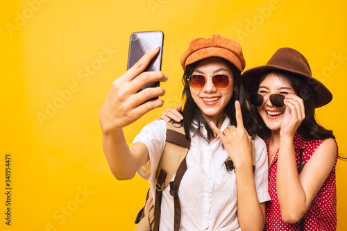 Two Asian pretty young girl smiling wear sunglasses and take a selfie by smartphone together, Young women backpacker take a selfie in studio yellow background.