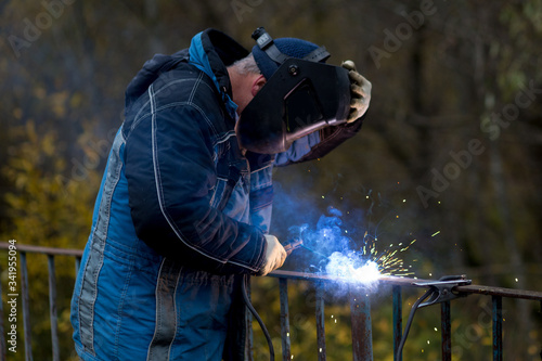 A man in a blue jacket is engaged in welding. A welder in a black mask repairs an iron fence. A man at work.