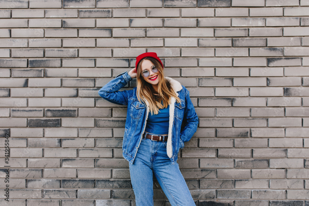 Happy girl in denim outfit standing in front of brick wall. Outdoor photo of caucasian young lady wears jeans and red hat expressing positive emotions.
