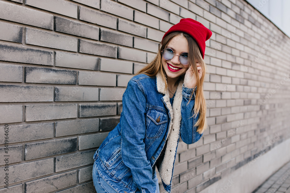 Ecstatic caucasian girl in denim attire and blue glasses posing with cute smile. Pleased young woman in red hat fooling around during street photoshoot. Ecstatic caucasian girl in denim attire and blue glasses posing with cute smile. Pleased young woman in red hat fooling around during street photoshoot.