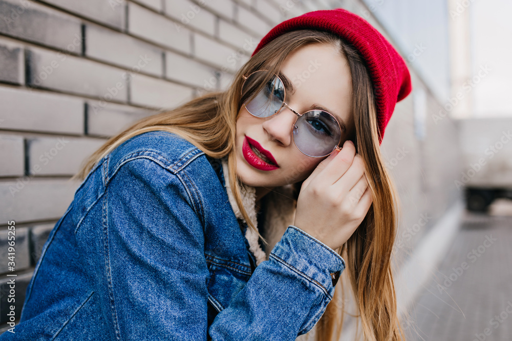 Winsome young woman with pale skin posing near brick wall with serious face expression. Outdoor photo of fashionable girl in trendy red hat chilling on the street. Winsome young woman with pale skin posing near brick wall with serious face expression. Outdoor photo of fashionable girl in trendy red hat chilling on the street.