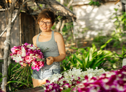 Beautiful young woman florist with glasses in tropical greenery makes a bouquet of purple orchids.