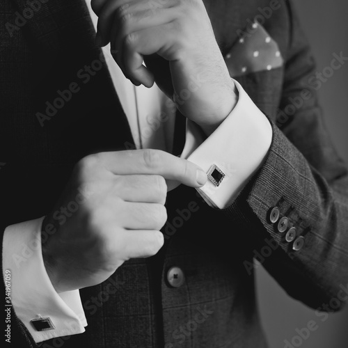 Elegant fashion man looking at his cufflinks while fixing them. Black and white photo of male hands. Handsome groom dressed in black formal suit, white shirt and tie is getting ready for wedding.
