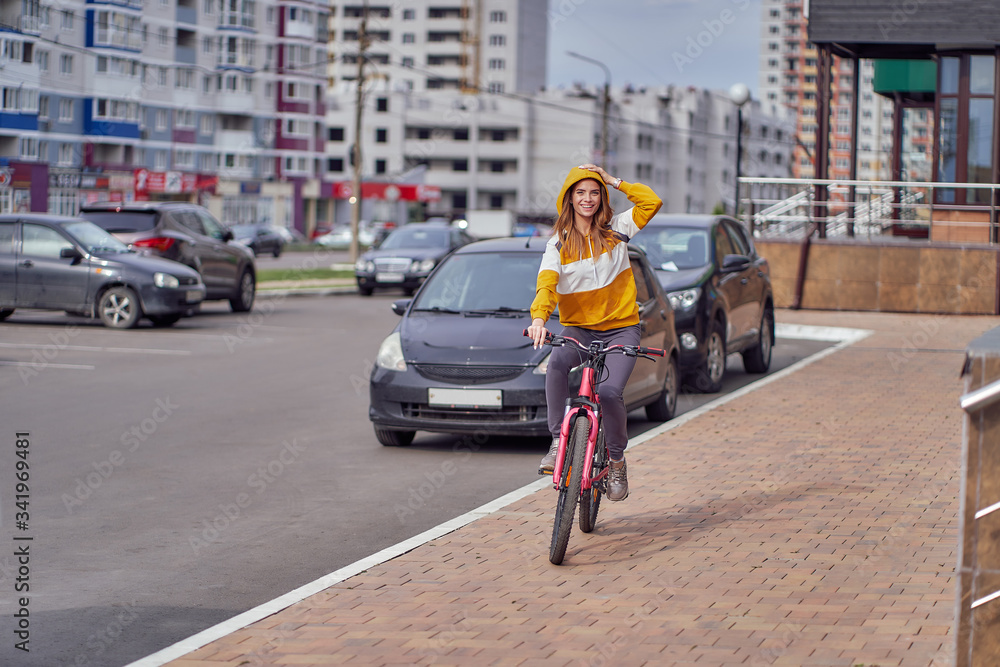 A girl rides a bicycle on the sidewalk in a city block in a bright yellow jacket.
