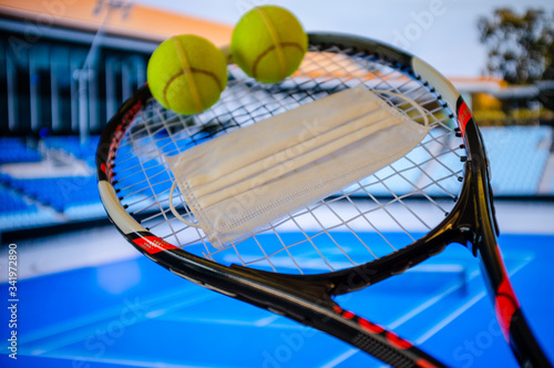 Tennis racket with a surgical mask and tennis balls, in front of a court background, referring cancellations due to coronavirus pandemic © gokercy