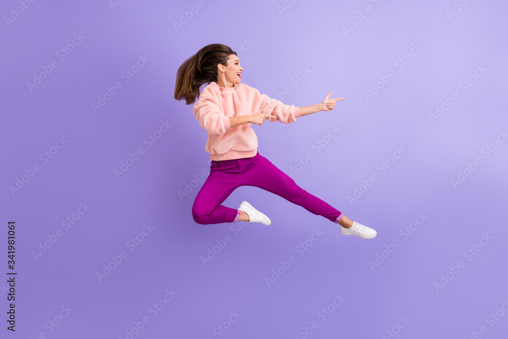 Full length profile photo of cheerful lady jumping high indicating fingers empty space amazing sale wear casual fluffy pullover pants shoes isolated purple color background Full length profile photo of cheerful lady jumping high indicating fingers empty space amazing sale wear casual fluffy pullover pants shoes isolated purple color background