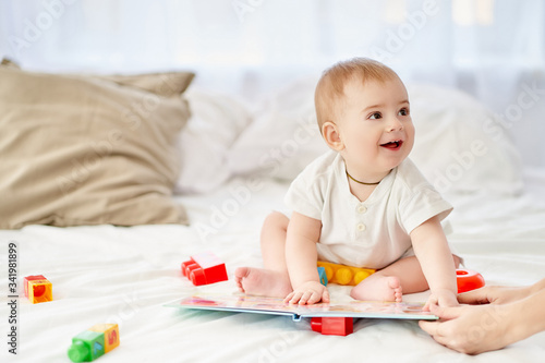 Cute happy baby reading a children's book on a white soft bed