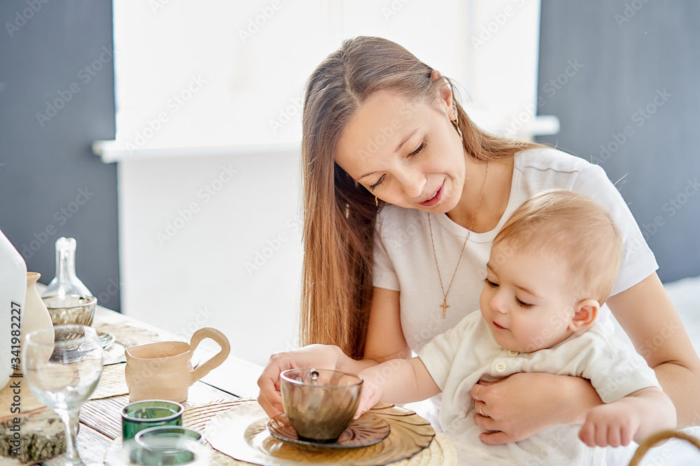 A young mother with a child on her knees drinks tea in a bright room.