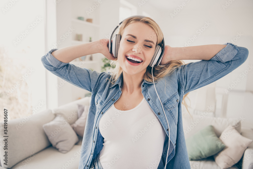 Naklejka premium Photo of pretty crazy blond lady rejoicing house party dancing near couch stay home listen cool earphones hold hands on ears eyes closed overjoyed quarantine time living room indoors