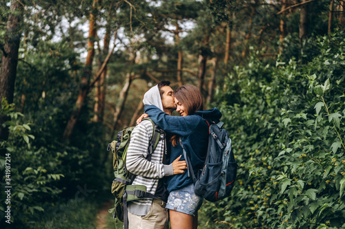 Young couple with backpacks on their backs in the forest. Loving man kisses his beautiful girlfriend on a hike in the woods