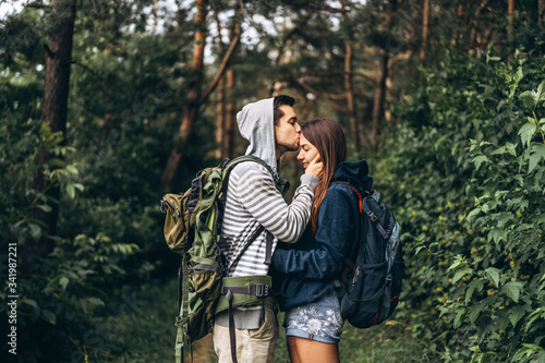 Young couple with backpacks on their backs in the forest. Loving man kisses his beautiful girlfriend on a hike in the woods