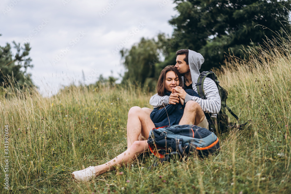 Woman with long hair and man sitting on the grass in the forest with backpacks. Have fun in nature, hugging and kissing Woman with long hair and man sitting on the grass in the forest with backpacks. Have fun in nature, hugging and kissing