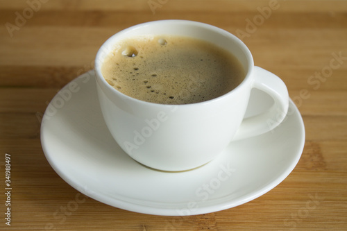A small white Cup of fresh espresso with foam and a saucer close up on a brown wooden background