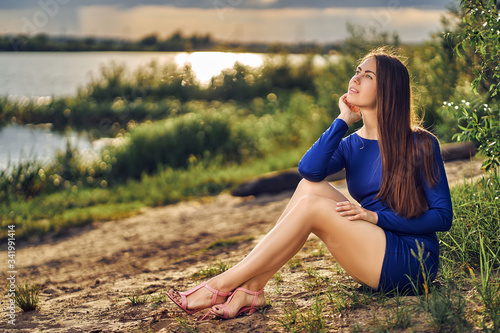 A young woman with long hair and in a short blue dress sits on the sand by the water and looks up. Summer outdoor recreation.