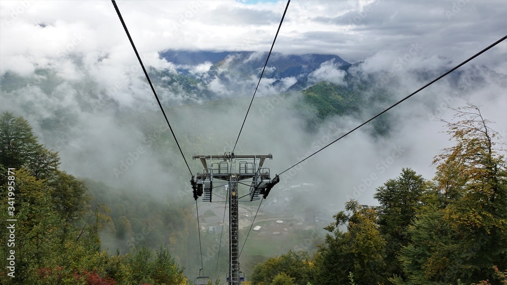 Cableway in the mountains of the Caucasus. Cabs fly right through very low white clouds. Visible mountains, yellow autumn leaves on the trees.