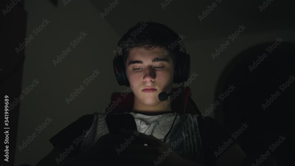 Young handsome man in headset with microphone using smartphone, sitting near computer at home late at night. Lighted face with a smart phone Young handsome man in headset with microphone using smartphone, sitting near computer at home late at night. Lighted face with a smart phone