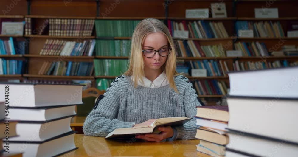 The girl is studying among many books in the reading room in the library. Beautiful girl with glasses. 4K