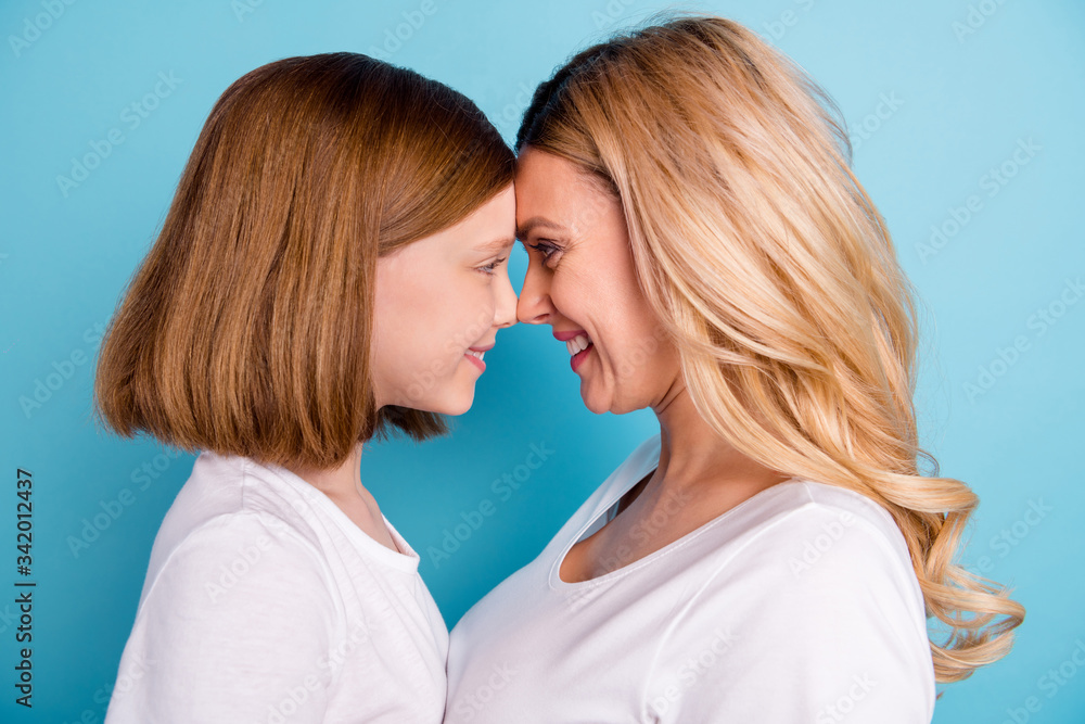 Cloeup profile photo of two people beautiful mommy lady small little daughter blonds look eyes leaning head to head wear casual white s-shirts isolated blue color background Cloeup profile photo of two people beautiful mommy lady small little daughter blonds look eyes leaning head to head wear casual white s-shirts isolated blue color background