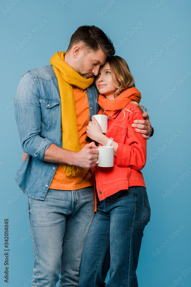 diseased couple in scarves hugging and holding cups with hot drinks isolated on blue diseased couple in scarves hugging and holding cups with hot drinks isolated on blue