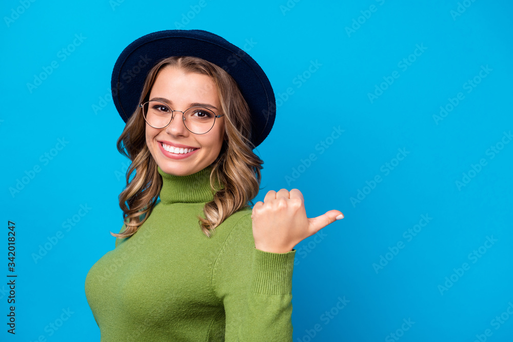 Photo of cheerful curly lady direct thumb finger side empty space advising cool shopping prices wear specs hat casual green turtleneck isolated blue color background Photo of cheerful curly lady direct thumb finger side empty space advising cool shopping prices wear specs hat casual green turtleneck isolated blue color background