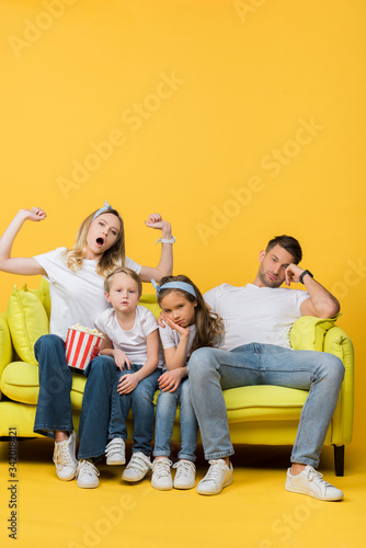 bored family yawning and watching movie on sofa with popcorn bucket on yellow