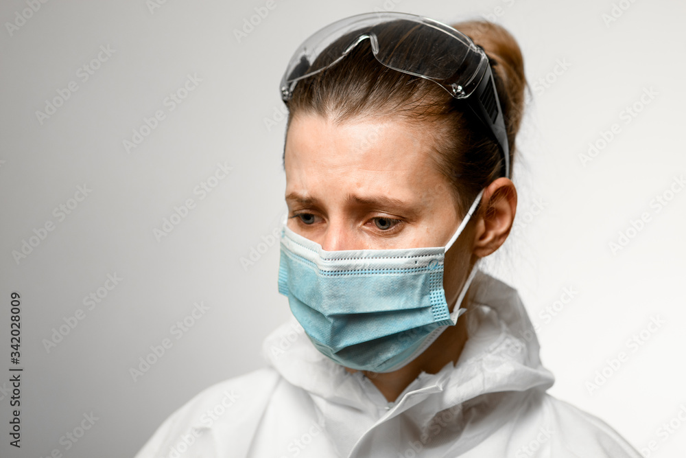 Young female nurse with goggles on her head and in medical mask with sad expression look down Young female nurse with goggles on her head and in medical mask with sad expression look down