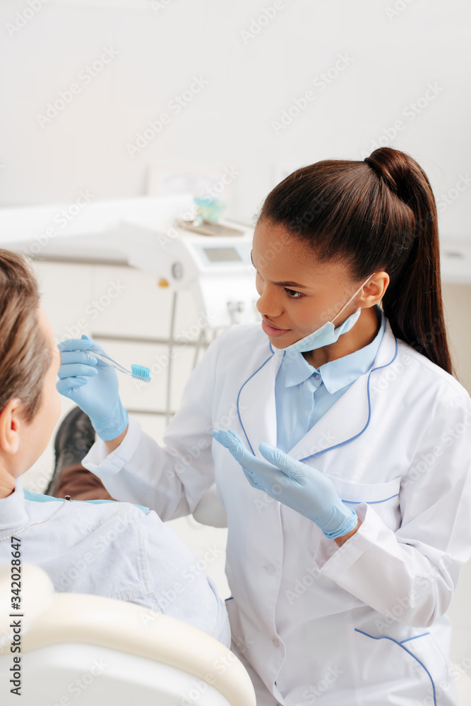 selective focus of african american dentist in latex gloves pointing with hand at toothbrush near patient selective focus of african american dentist in latex gloves pointing with hand at toothbrush near patient