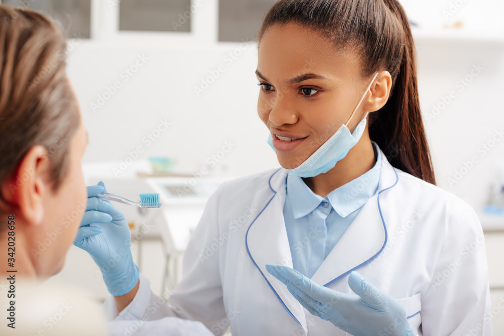 selective focus of happy african american dentist in latex gloves pointing with hand at toothbrush near patient selective focus of happy african american dentist in latex gloves pointing with hand at toothbrush near patient