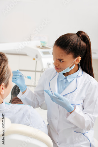 selective focus of african american dentist in latex gloves pointing with hand at toothbrush near patient