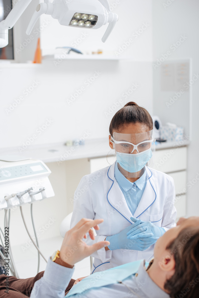 selective focus of african american dentist in medical mask and face shield near man in clinic