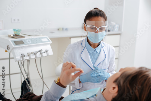selective focus of african american dentist in medical mask and face shield near patient in clinic