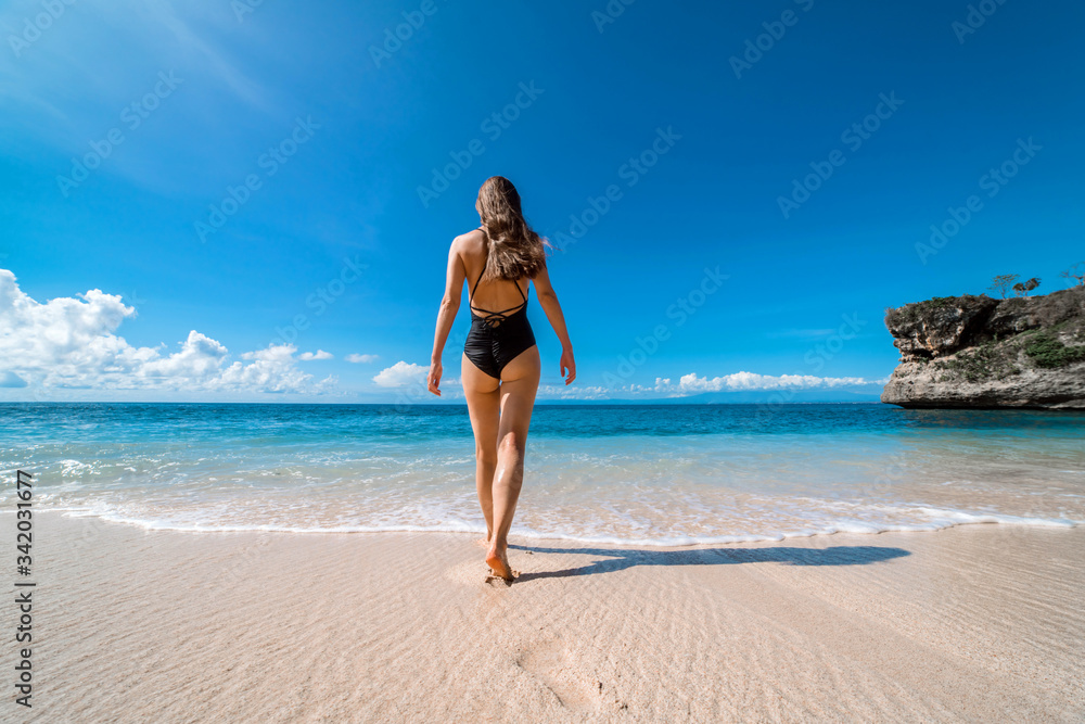 Beautiful young tourist girl walking on an empty beach in Bali. A sexy girl in a black swimsuit is photographed from behind walking barefoot on the sand towards the ocean.