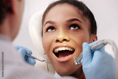 selective focus of dentist in latex gloves holding dental instruments near happy african american woman with opened mouth