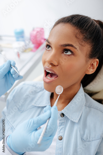 dentist in latex gloves holding dental equipment near african american patient with opened mouth