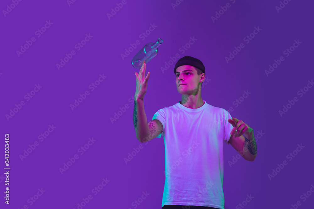 Throws up a bottle of water. Caucasian man's portrait isolated on purple studio background in pink neon light. Beautiful male model in casual. Concept of human emotions, facial expression, sales, ad. Throws up a bottle of water. Caucasian man's portrait isolated on purple studio background in pink neon light. Beautiful male model in casual. Concept of human emotions, facial expression, sales, ad.